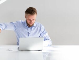 Ein Mann sitzt an seinem Laptop und hält einen Stapel Ordner weg von sich