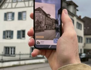Eine Person hält ein Smartphone hoch, das ein altes Foto derselben Straßenszene zeigt, die im Hintergrund zu sehen ist. Im Hintergrund sind traditionelle Gebäude mit Fachwerk und ein Torbogen zu sehen. Der Himmel ist bewölkt.