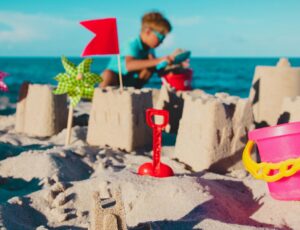 Kind baut Sandburgen am Strand. Im Vordergrund ist ein pinkfarbener Eimer.
