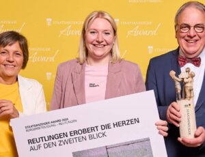 Drei Personen stehen vor einem gelben Hintergrund mit dem Schriftzug „Staatsanzeiger Award“. Eine Frau hält eine Urkunde, eine zweite Frau lächelt in der Mitte, und ein Mann hält eine Trophäe in der Form einer kleinen Skulptur. Sie feiern eine Auszeichnung mit dem Titel: „Reutlingen erobert die Herzen auf den zweiten Blick“.