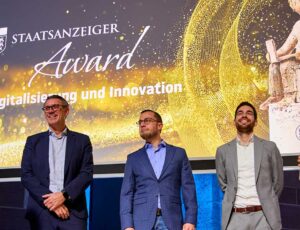 Eine Bühne während einer Preisverleihung. Im Hintergrund steht auf einer großen Leinwand „Staatsanzeiger Award – Digitalisierung und Innovation“ mit einem goldenen Design. Vier Männer stehen vorne und lächeln, während einer eine Trophäe hält. Ein Moderator auf der linken Seite spricht zu den Preisträgern.