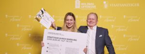 Ein Mann und eine Frau posieren vor einer gelben Wand mit den Logos „Holzbau Offensive“ und „Staatsanzeiger Award“. Die Frau hält ein großes Plakat mit dem Titel „Stadttour durch Marburg wird zum Hit auf TikTok“ sowie eine Urkunde, während der Mann stolz in die Kamera blickt.
