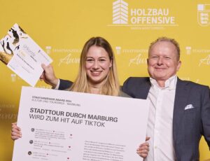 Ein Mann und eine Frau posieren vor einer gelben Wand mit den Logos „Holzbau Offensive“ und „Staatsanzeiger Award“. Die Frau hält ein großes Plakat mit dem Titel „Stadttour durch Marburg wird zum Hit auf TikTok“ sowie eine Urkunde, während der Mann stolz in die Kamera blickt.