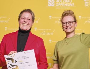 Zwei Frauen posieren vor einer gelben Wand mit den Logos „Holzbau Offensive“ und „Staatsanzeiger Award“. Die Frau links hält eine Urkunde, und die Frau rechts hebt triumphierend die Faust. Beide lächeln und strahlen Freude aus.