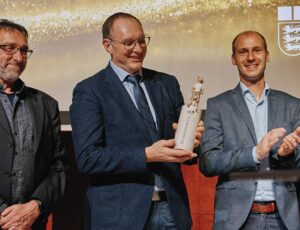 Eine Gruppe von Personen steht auf einer Bühne vor einer Leinwand mit dem Staatsanzeiger-Logo. Zwei Männer in der Mitte präsentieren eine Trophäe, während die anderen applaudieren und lächeln. Die Szene zeigt eine Preisverleihung im Rahmen des Staatsanzeiger Awards.