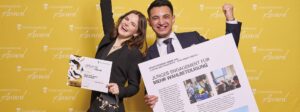 Eine junge Frau und ein junger Mann stehen vor einer gelben Wand mit dem Logo „Staatsanzeiger Award“. Die Frau hält eine Urkunde und jubelt, während der Mann ein großes Plakat mit der Überschrift „Junges Engagement für mehr Wahlbeteiligung“ präsentiert. Beide strahlen vor Freude.