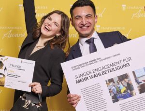 Eine junge Frau und ein junger Mann stehen vor einer gelben Wand mit dem Logo „Staatsanzeiger Award“. Die Frau hält eine Urkunde und jubelt, während der Mann ein großes Plakat mit der Überschrift „Junges Engagement für mehr Wahlbeteiligung“ präsentiert. Beide strahlen vor Freude.