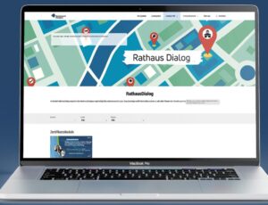 Ein Laptop auf einem blauen Hintergrund zeigt eine Website mit dem Titel „Rathaus Dialog“. Auf der Seite sind eine grafische Karte mit Markierungen und Informationen zum Thema digitale Verwaltung zu sehen. Die Darstellung vermittelt ein modernes, digitales Konzept.