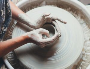 Eine Nahaufnahme von Händen, die einen Tonklumpen auf einer rotierenden Töpferscheibe formen. Die Szene zeigt die kreative Arbeit eines Keramikers in einem Atelier, umgeben von Werkzeugen und Ton.