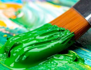 Ein Pinsel mit frischer, satter grüner Farbe streicht über eine bunte Farbpalette mit gelben, blauen und orangen Tönen. Die Szene symbolisiert Kreativität und Nachhaltigkeit.