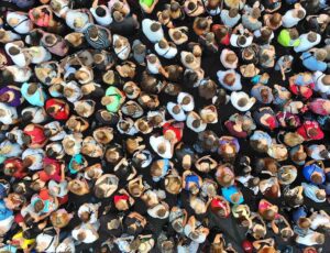Luftaufnahme einer großen Menschenmenge, dicht gedrängt auf einer dunklen Fläche, mit unterschiedlich farbiger Kleidung aus der Vogelperspektive.
