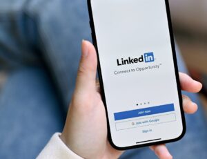 Hand hält ein Smartphone mit dem LinkedIn‑Startbildschirm; rechts ist eine Hand mit einem Pappbecher zu sehen, im Hintergrund unscharfe Jeansbekleidung.