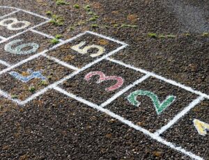 Asphaltboden mit bunter Hüpfekästchen‑Grafik in Kreide; die Felder sind von 1 bis 8 nummeriert, einige Zahlen sind farbig ausgefüllt, im Hintergrund kleine Grasbüschel auf dem Weg.