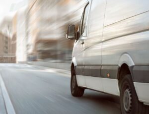 Weißer Kleinbus fährt mit Bewegungsunschärfe durch eine Stadtstraße, im Vordergrund ist das Heck mit rotem Rücklicht und die Seite des Fahrzeugs zu sehen.