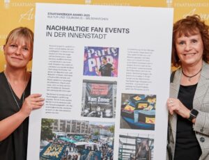 Zwei Vertreterinnen zeigen ein Plakat zu nachhaltigen Fan Events in der Innenstadt von Gelsenkirchen